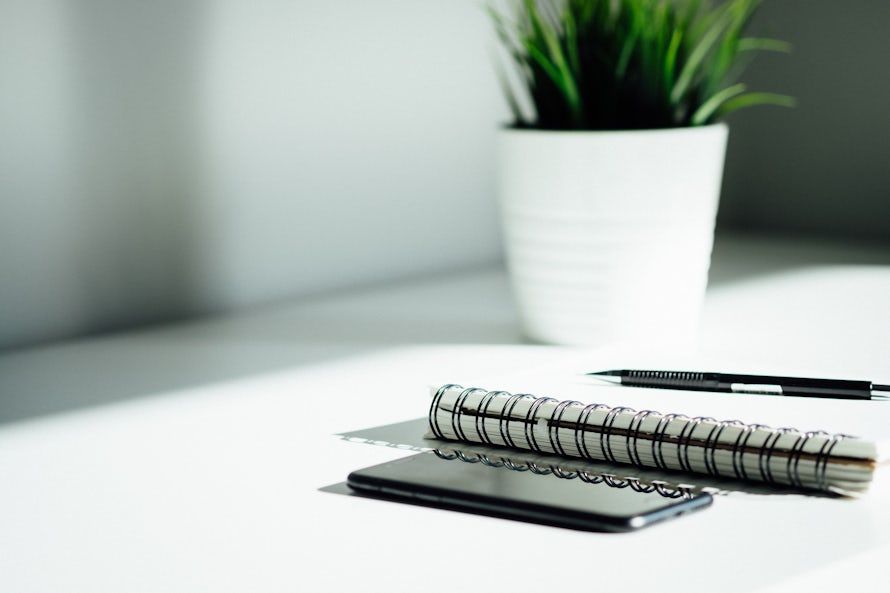 A notepad, mechanical pencil, phone and plant on very brightly lit desk